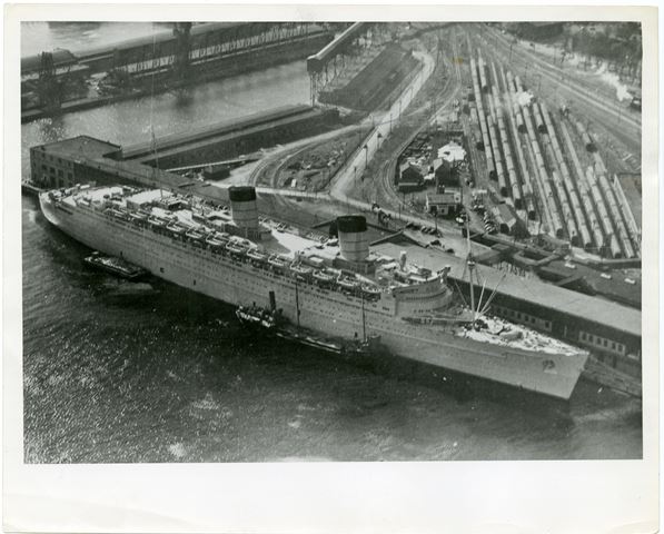 Vu du ciel, un grand navire de guerre peint en gris accoste à une série de hangars bas au bord de l’eau, avec des gares de triage et un deuxième hangar en arrière. Des remorqueurs aident le navire à s’amarrer. À gauche de l’image et à l’arrière-plan se trouvent certaines des structures de manutention des céréales et des marchandises pour les quais voisins.