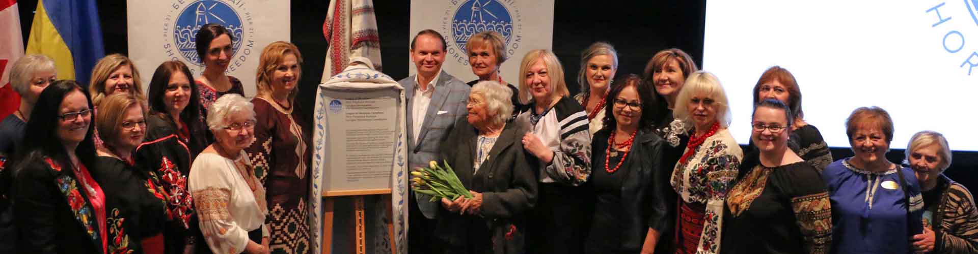 A group of 20 people, almost all women, some in traditional Ukrainian costumen, pose with a plaque on an easel that's just been unveiled.
