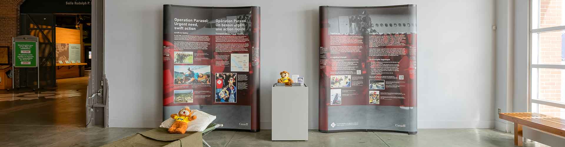 Deux grands stands avec le texte et les images de l'exposition. Deux ours Zeddy sont présentés : l'un sur un lit d'enfant et l'autre sur un support.