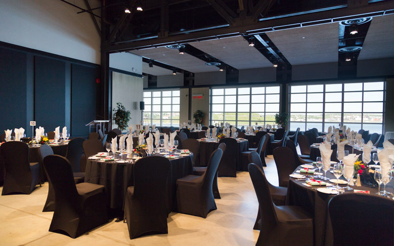 Une salle dont les fenêtres allant du sol au plafond offrent une vue sur le port et l’île et des tables de banquet ovales avec des chaises et des ustensiles de cuisine.