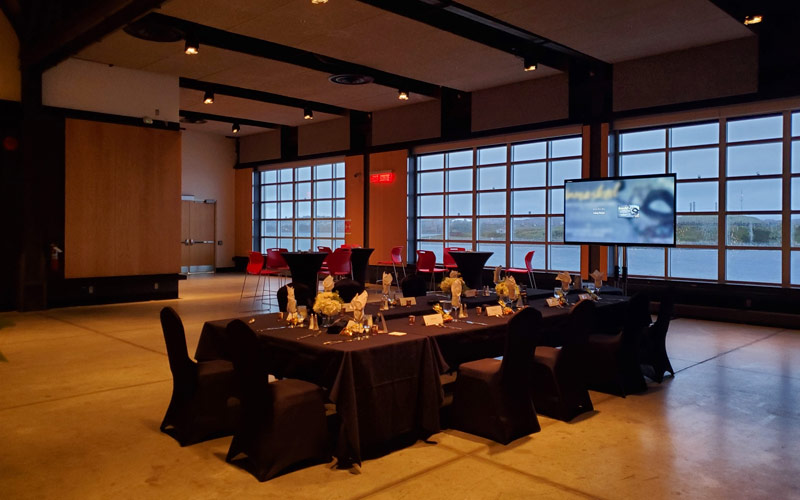 Des tables de banquet et des chaises disposées en forme de U offrant une vue sur le port par les fenêtres. Un téléviseur est installé pour une présentation.