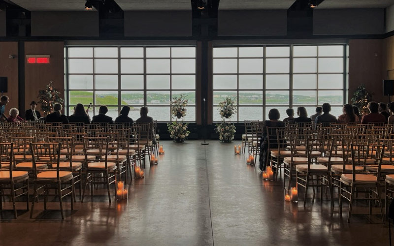 Des chaises blanches faisant face à des fenêtres offrant une vue sur le port sont disposées dans un style salle de spectacle en vue d’une cérémonie de mariage. Deux supports floraux et un pied de micro se trouvent au bout de l’allée. Des bougies dans des vases cylindriques sont placées à côté des chaises, le long de l’allée.