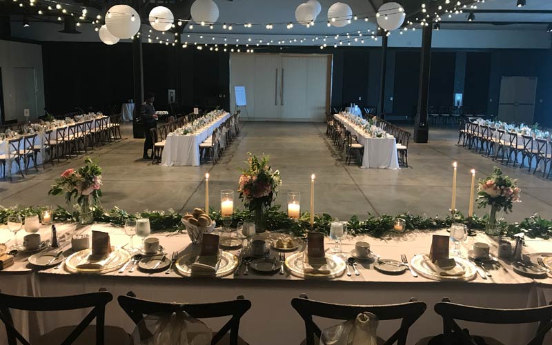 Une salle aménagée en vue d’un mariage et offrant une longue table d’honneur sur une estrade, décorée de guirlandes, de bougies et de bouquets. Des tables sont disposées en quatre longues rangées, des chaises en bois étant placées de chaque côté.