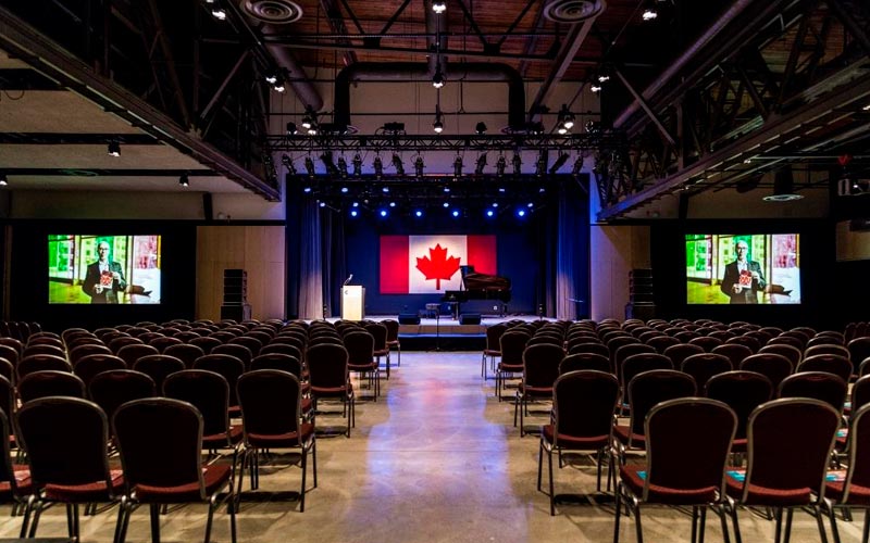 Une salle préparée en vue d’une présentation et d’un spectacle, des chaises disposées dans un style salle de spectacle et une allée centrale. Sur la scène se trouvent un podium, un piano à queue noir et une toile de fond représentant le drapeau canadien. Deux toiles de projection sont disposées de part et d’autre de la scène.