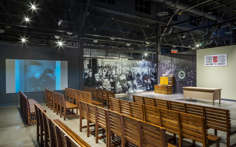 Une exposition du Musée offrant de longs bancs en bois faisant face à une table et à un mur sur lequel se trouve un panneau « Bienvenue au Canada ». À gauche des bancs se trouvent une toile de projection et une image historique d’immigrants.