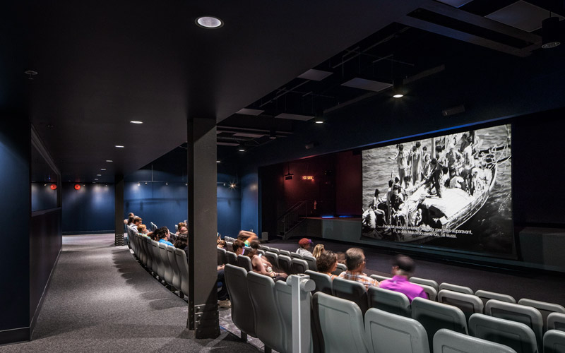 Un amphithéâtre avec une centaine de chaises où se trouve une douzaine d’invités assis face à un écran déroulant sur lequel est projetée l’image en noir et blanc d’un navire.
