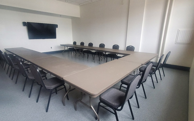 Une petite pièce aux hauts plafonds pouvant accueillir quinze invités, équipée de tables rectangulaires et de chaises de banquet disposées en forme de U. L’installation fait face à un téléviseur en vue d’une présentation.