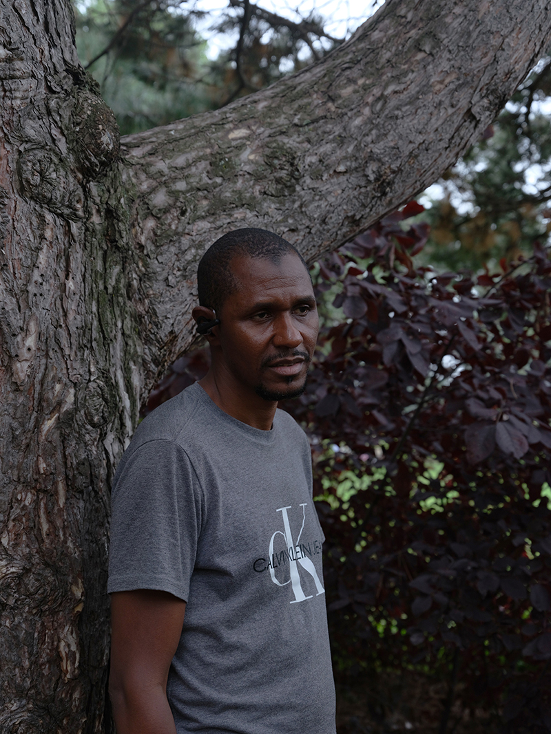 Un homme en T-shirt gris s’appuie sur un grand arbre.