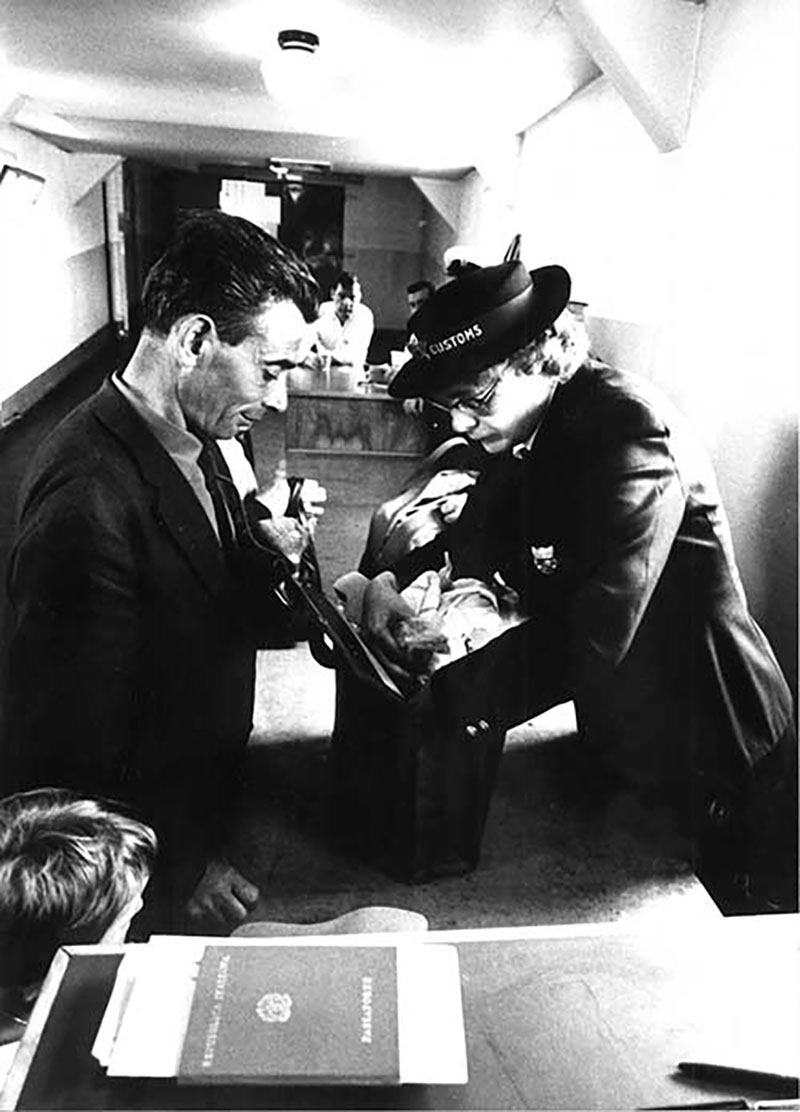 Une douanière inspecte des bagages tandis que le propriétaire observe.