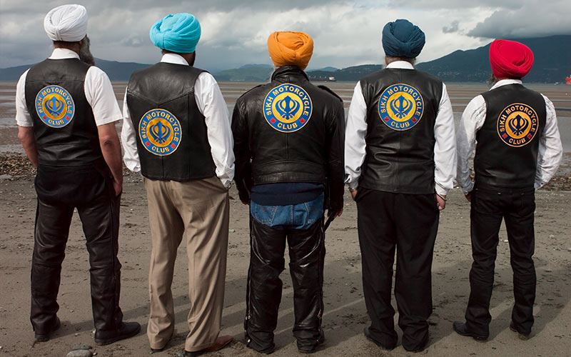 Six hommes se tiennent dos à la caméra, portant des gilets arborant l’inscription « Sikh Motorcycle Club ».