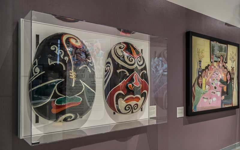 Deux grands masques dans une vitrine à côté d’un tableau représentant une famille à table, tous deux accrochés au mur.