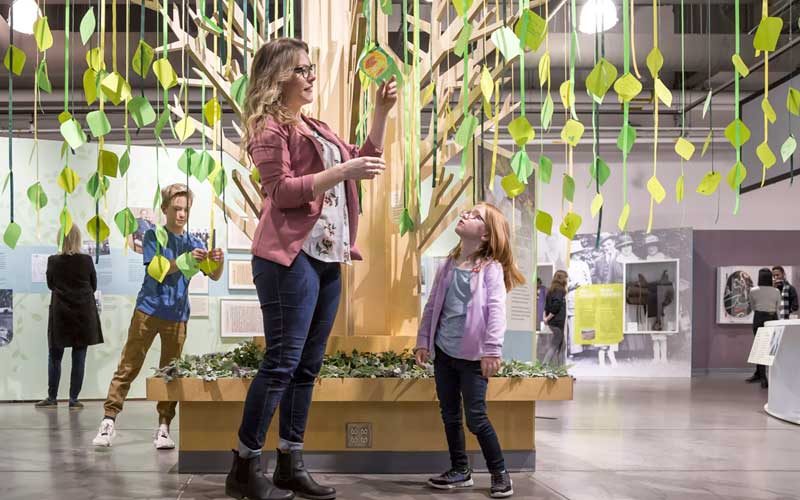 Une personne adulte et deux enfants se tiennent sous un arbre construit, lisant des notes sur des feuilles de papier.