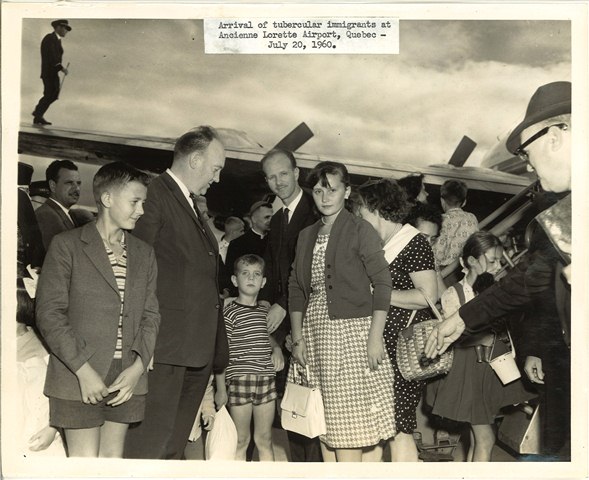 Photo en tons sépia d’un grand groupe de personnes à la peau claire habillées de manière formelle sur un tarmac avec un avion derrière eux.