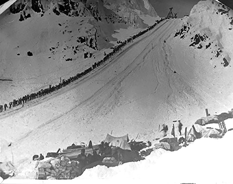 Des personnes formant une très longue ligne montent lentement sur une étendue enneigée.