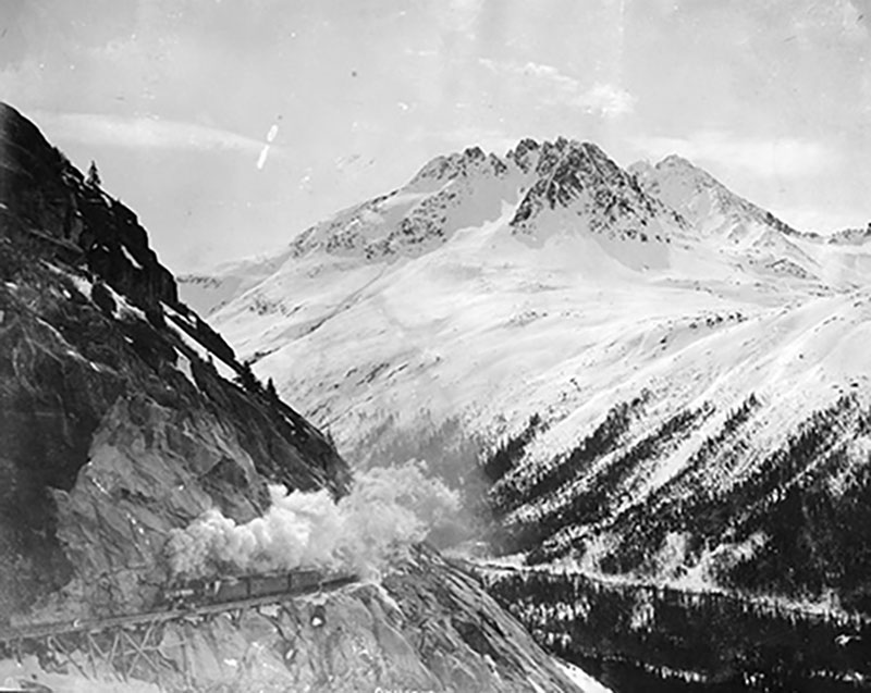 Un train avec une colonne de fumée avance le long d’un col de montagne.