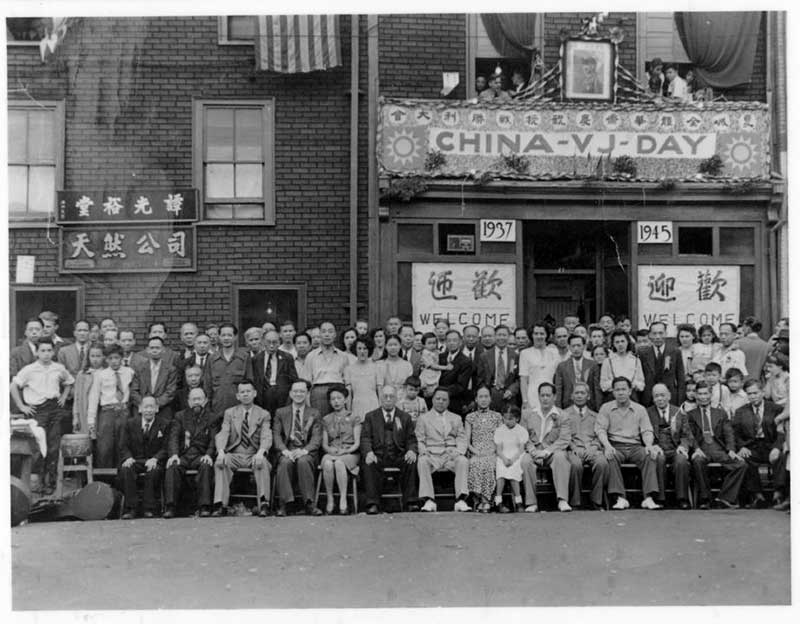 Un grand groupe de personnes pose à l’extérieur d’un bâtiment avec une banderole portant l’inscription « CHINA-VJ-DAY » (Jour de Victore sur le Japon de la Chine).