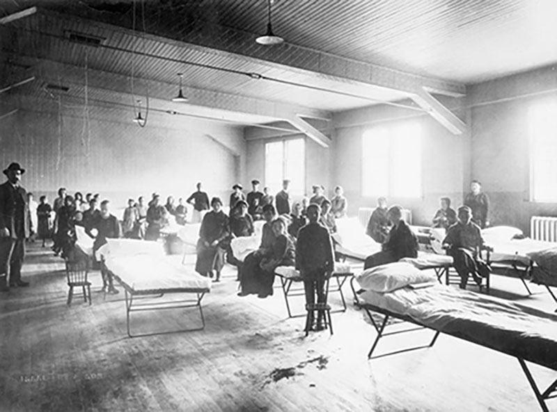 Un grand dortoir est rempli de lits de fortune; des gens y sont assis.