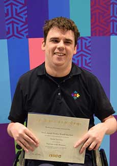 Un jeune homme en fauteuil roulant est titulaire d’un certificat, souriant. Le texte du certificat n’est pas lisible.