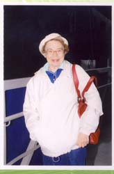 Ursula plus âgée, en veste blanche, chapeau et sac à main brun.