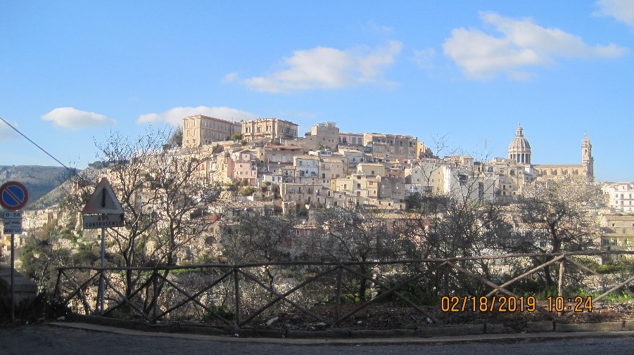 La ville d’Ibla vue de loin, avec une barrière et des arbres devant.