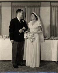 Photographie des époux le jour du mariage, devant une table de banquet.