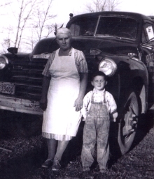 Femme en tablier et un petit garçon portant une salopette, appuyés contre une vieille voiture.