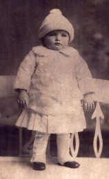 Bébé debout, portant des vêtements blancs et un bonnet tricoté blanc.